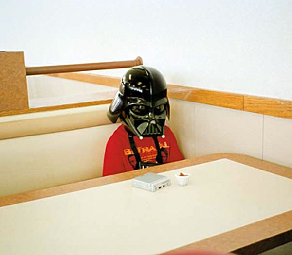 Sad-looking kid in a Darth Vader mask sitting alone at a fast-food restaurant table.