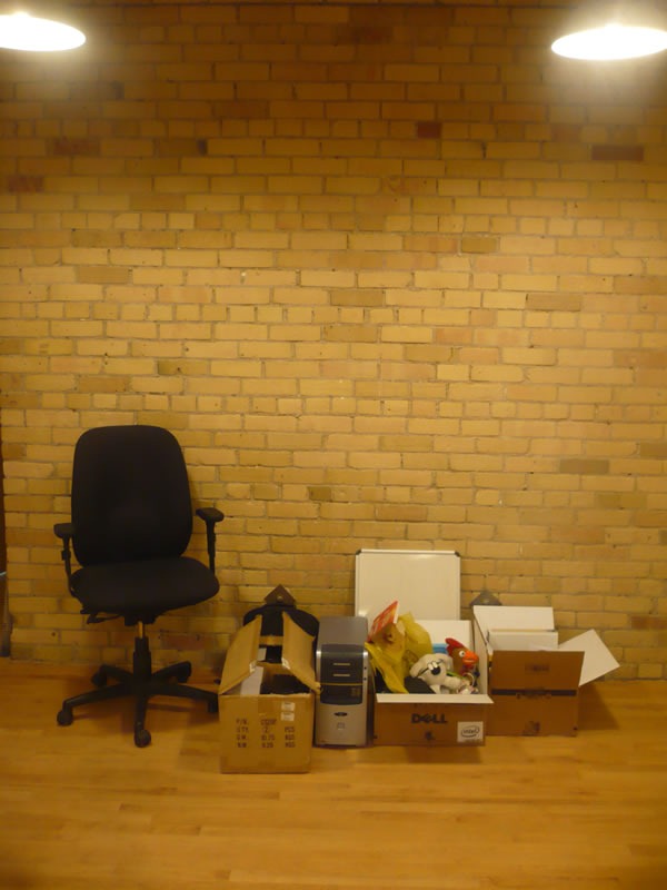 An office chair, a computer and some boxes lined up against an interior brick wall