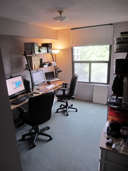 Carpeted apartment bedroom converted into a home office, showing a long desk with computers and a window looking out onto treetops