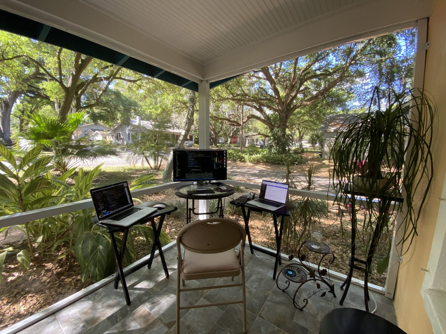 A screened-in front porch with a chair, table, and two laptop computers. There is a view of a garden with a tree-lined residential street corner.