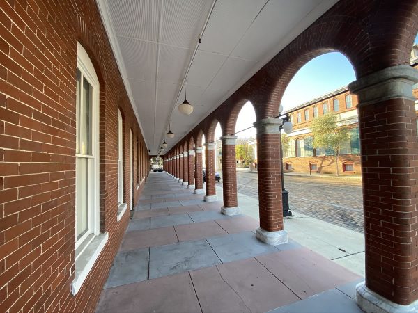 Photo: A long shot of the sidewalk in front of The Undercroft. The Undercroft is a red brick building with white windows, and the walkway is a covered one with brick arches.