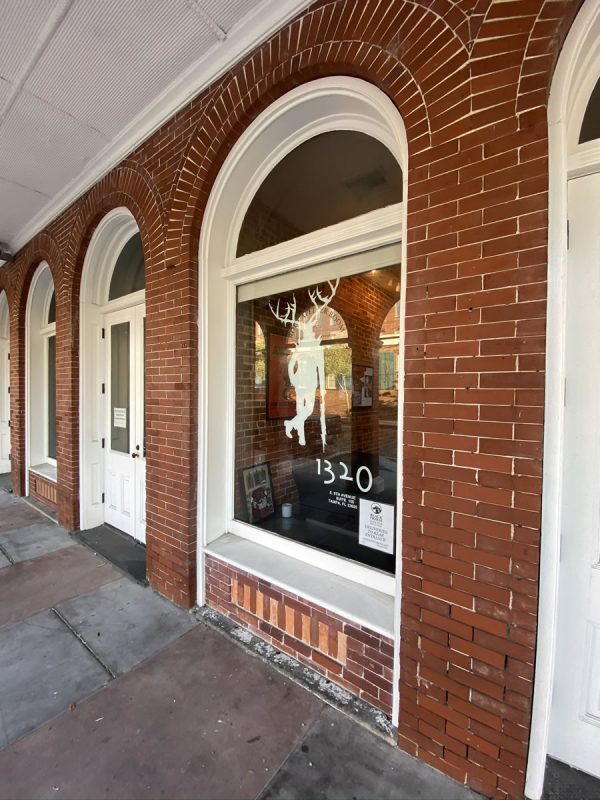 Photo: The front entrance of The Undercroft. On the left is a white wooden double door in an arched doorway with large glass windows. To the right is a large glass window featuring The Undercroft’s mascot (a stag standing like a human leaning against an umbrella) and the number 1320 (the address).