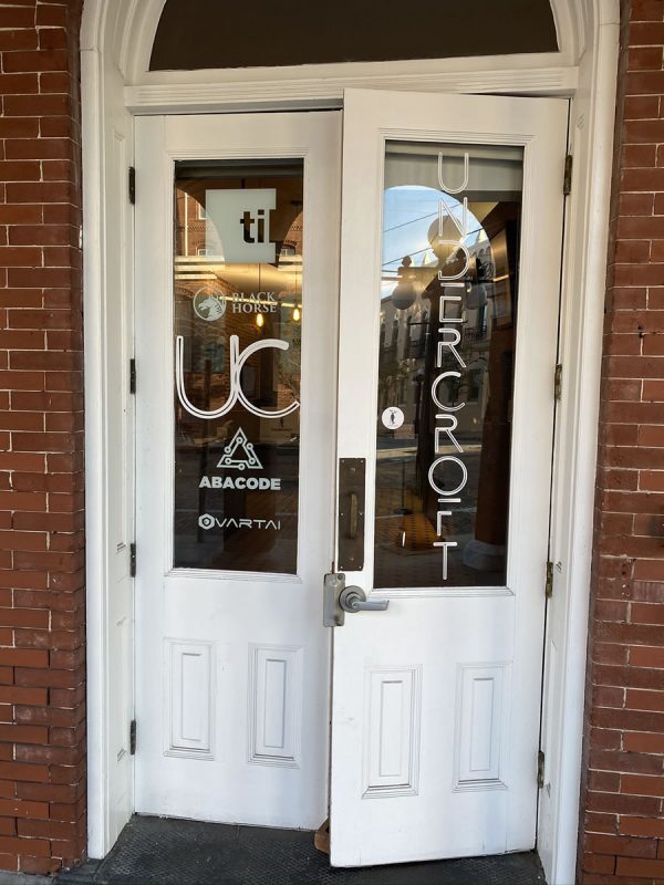 Photo: The front door of The Undercroft. Two white wooden double doors in an arched doorway. The Windows on the doors bear the logos of The Undercroft, til, Black Horse, UC, Abacode, and Vartai.