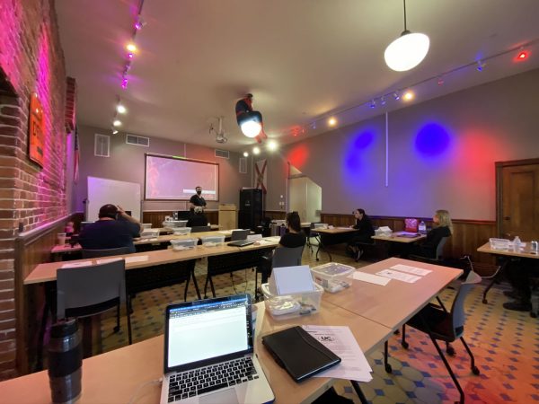 Photo: The Undercroft’s classroom. A room with an exposed brick wall on one side, many large table-sized wooden desks, red, white and blue spotlights and a large projection screen at the front. A Spider-Man mannequin sits atop one of the ceiling lights. A MacBook Pro is in the foreground.