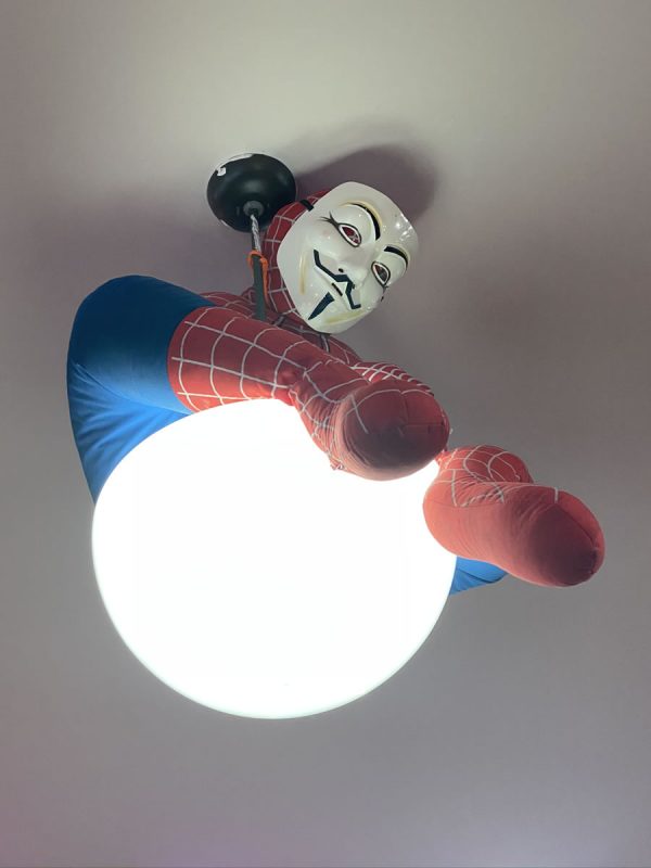 Photo: Close-up shot of the ceiling light in the center of The Undercroft’s classroom, which has a Spider-Man mannequin perched atop it. The mannequin is wearing a Guy Fawkes mask.