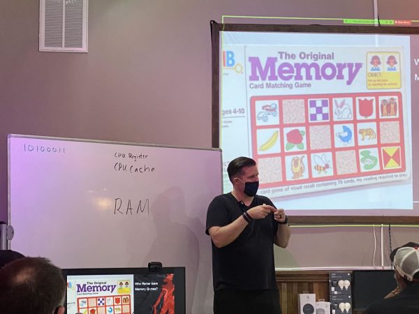 Photo: The instructor, Tremere, delivers a presentation. Behind him to the left as a whiteboard with “10100011”, “CPU register”, “CPU cache”, and “RAM” written on it. Behind and above him to the right is the projection screen, with a photo of the old board game “Memory” on display.