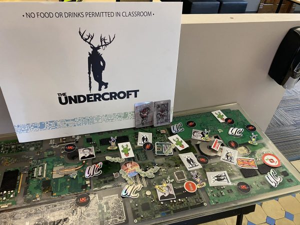 Photo: The swag table in The Undercroft’s lobby, which has a number of printed circuit boards embedded in it. The table has an assortment of Undercroft and security stickers on it, and at the back in a sign bearing The Undercroft’s name and mascot.