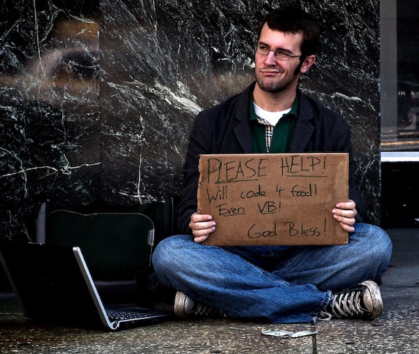 Programmer sitting on sidewalk with laptop holding up sign made from a cardboard box that says “Please help! Will code 4 food! Even VB! God bless!”