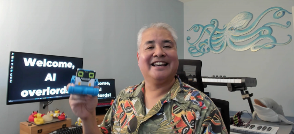 Joey deVilla, seen in his home office, holding a small robot in his right hand. The text “Welcome, AI overloads!” appears on screens in the background.