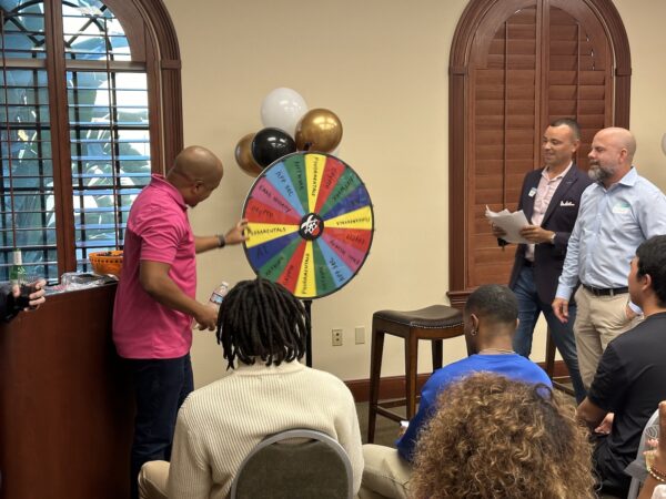 A contestant spins the multi-colored Wheel of Misfortune and Jason Allen and Jonas Kelley look on.