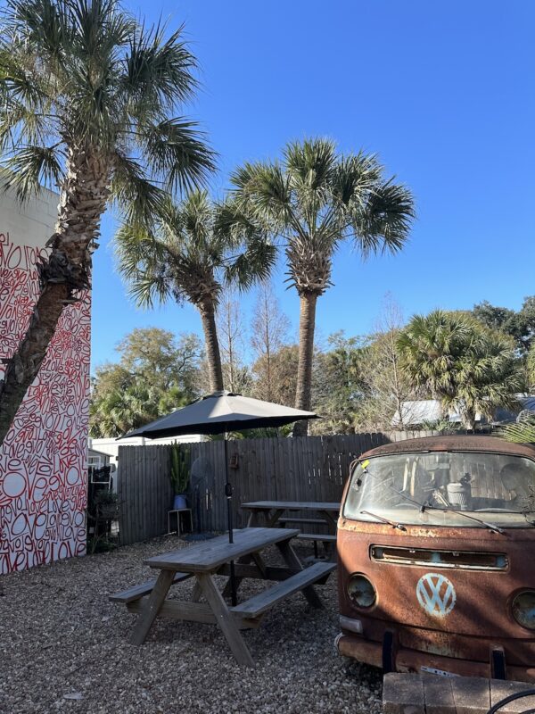 The courtyard at Spaddy’s Coffee in Seminole Heights, Tampa. It’s an open space sandwiched between two one-storey buildings with picnic and patio tables, and palm trees. The sky is blue and cloudless. There is a rusted-out VW van in the foreground.