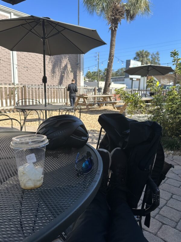 My view of Spaddy’s Coffee from my table. My legs are stretched out and my feet are resting on another chair. A finished coffee, my sunglasses, and  my bike helmet are resting on the table. The courtyard is in the background.