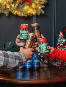 Arms in Christmas sweaters toasting with cans of beer under a Christmas wreath